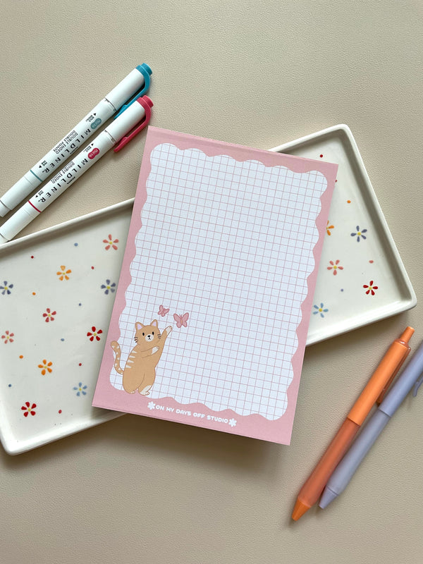 Grid notepad with a cute orange cat playing with butterflies in the bottom left corner. This notepad also has a pink wavy boarder. 