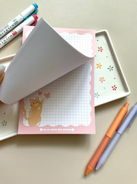 Grid notepad with a cute orange cat playing with butterflies in the bottom left corner. This notepad also has a pink wavy boarder. 