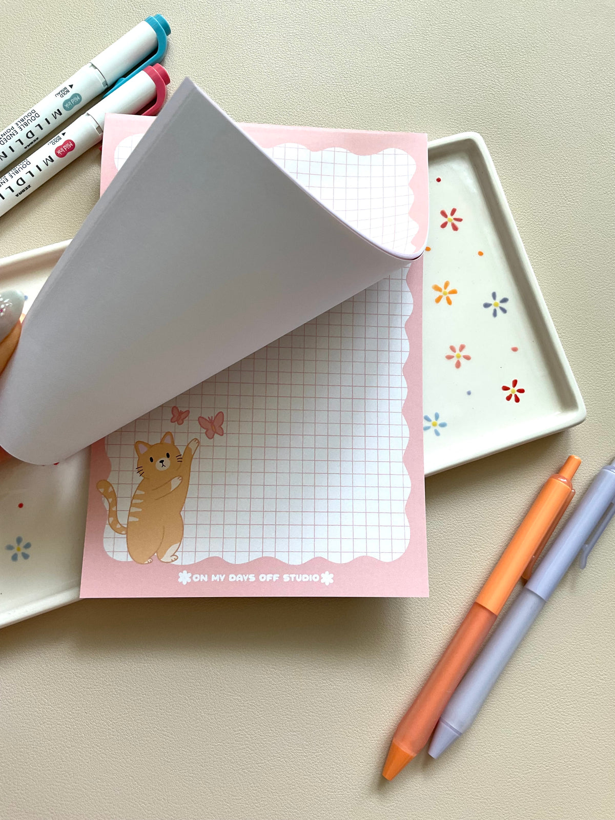Grid notepad with a cute orange cat playing with butterflies in the bottom left corner. This notepad also has a pink wavy boarder. 