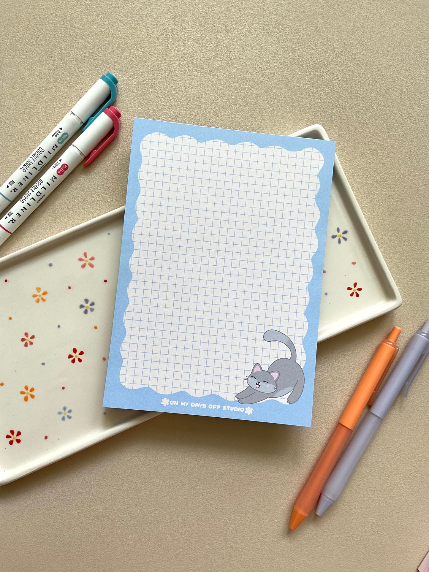 Grid notepad with a cute grey cat stretching in the bottom right corner. This notepad also has a blue wavy boarder. 