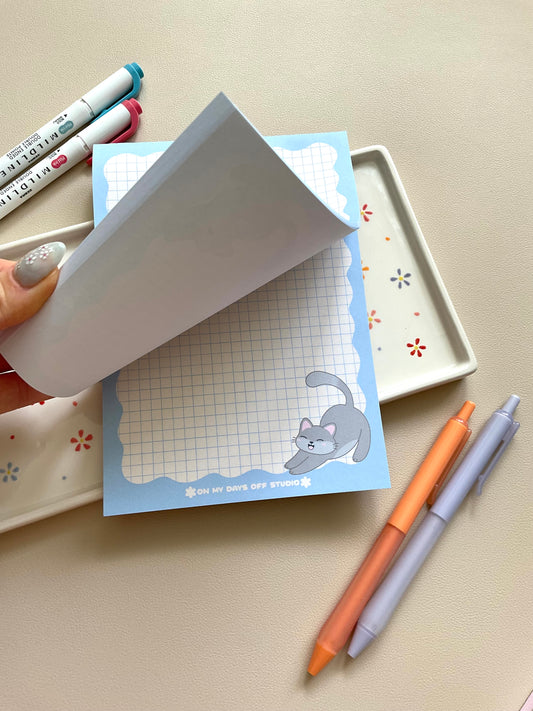 Grid notepad with a cute grey cat stretching in the bottom right corner. This notepad also has a blue wavy boarder. 