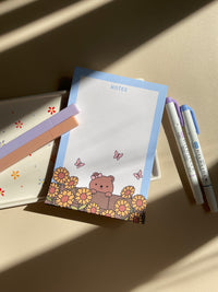 Cute notepad with a light blue boarder and beige background. A cute illustration of a bear peaking over a brick wall with sunflowers and butterflies