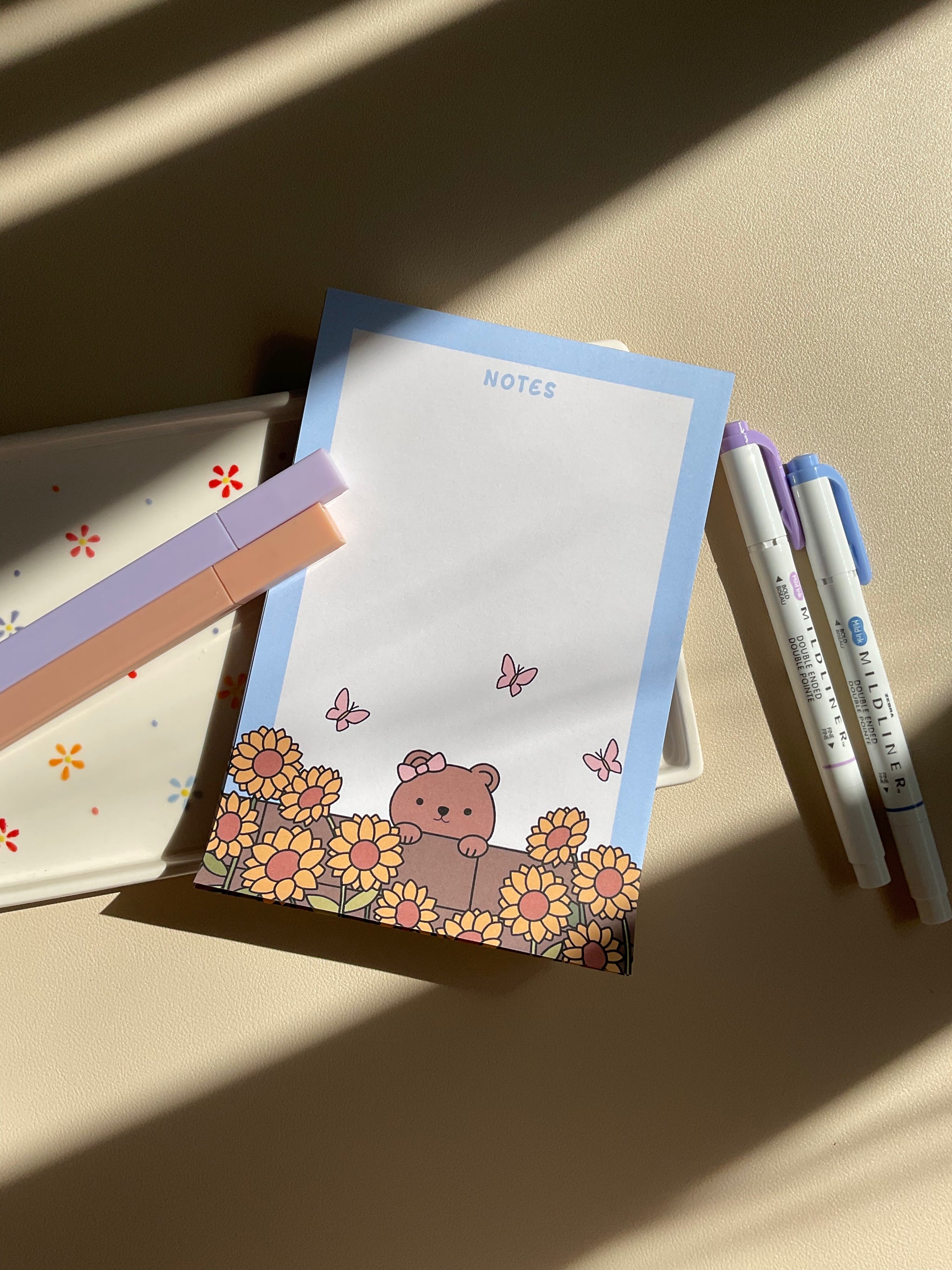 Cute notepad with a light blue boarder and beige background. A cute illustration of a bear peaking over a brick wall with sunflowers and butterflies