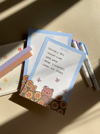 Cute notepad with a light blue boarder and beige background. A cute illustration of a bear peaking over a brick wall with sunflowers and butterflies