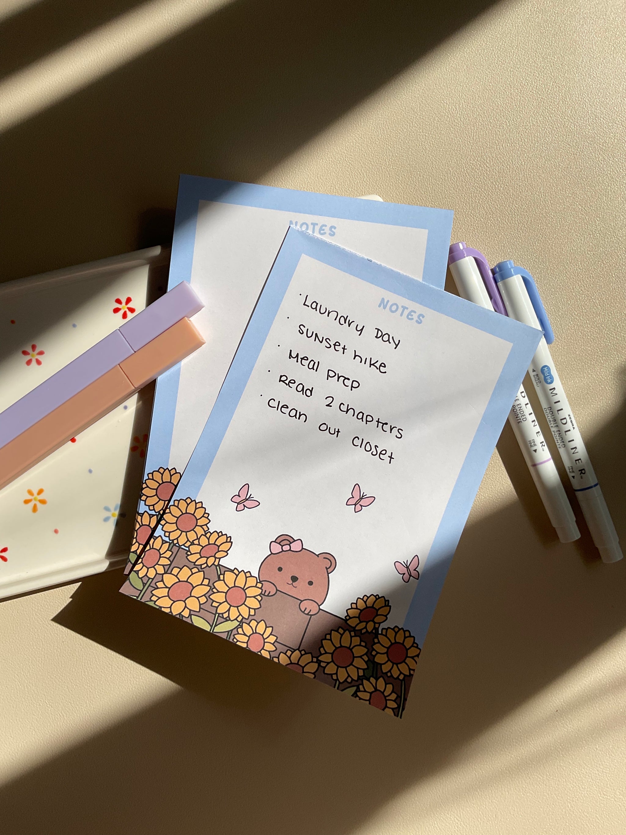 Cute notepad with a light blue boarder and beige background. A cute illustration of a bear peaking over a brick wall with sunflowers and butterflies