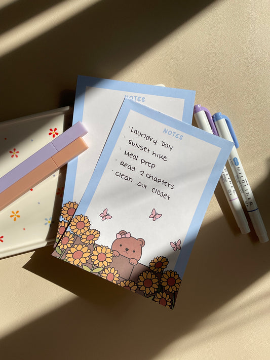 Cute notepad with a light blue boarder and beige background. A cute illustration of a bear peaking over a brick wall with sunflowers and butterflies