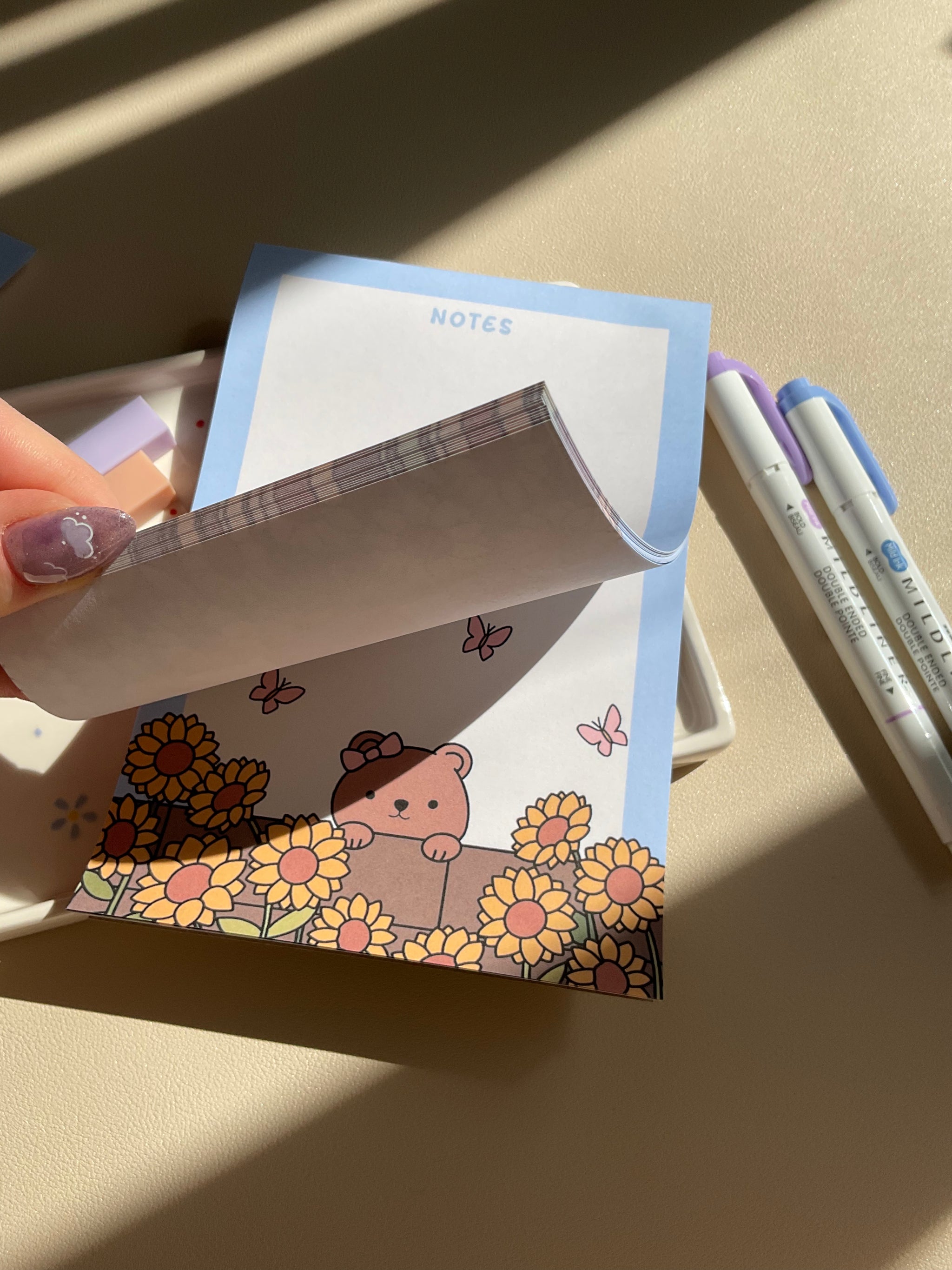 Cute notepad with a light blue boarder and beige background. A cute illustration of a bear peaking over a brick wall with sunflowers and butterflies