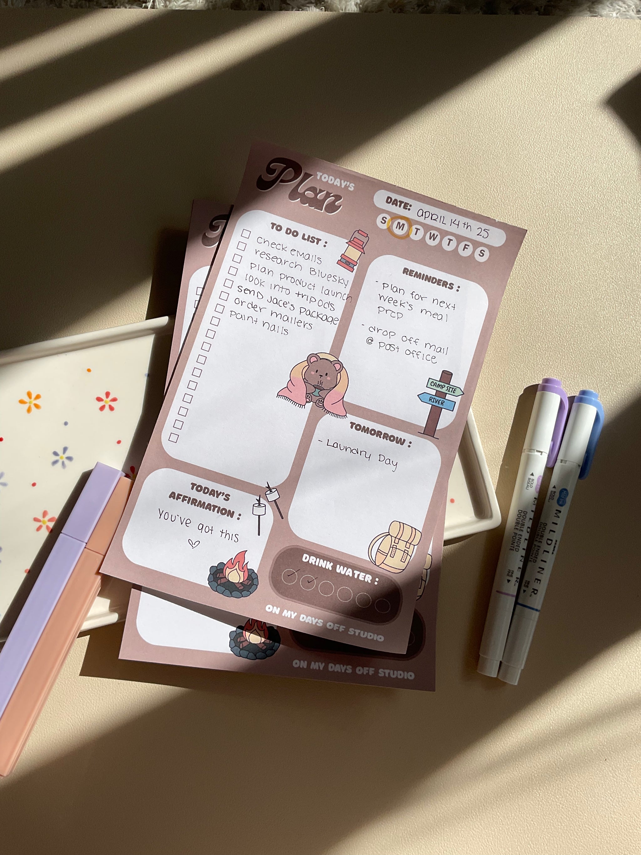 A daily planner notepad with a camp theme, featuring cute illustrations of a bear and camping related things. It has sections for a t do list, reminders, tomorrow list, today's affirmation and water in take tracker.