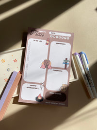 A daily planner notepad with a camp theme, featuring cute illustrations of a bear and camping related things. It has sections for a t do list, reminders, tomorrow list, today's affirmation and water in take tracker.