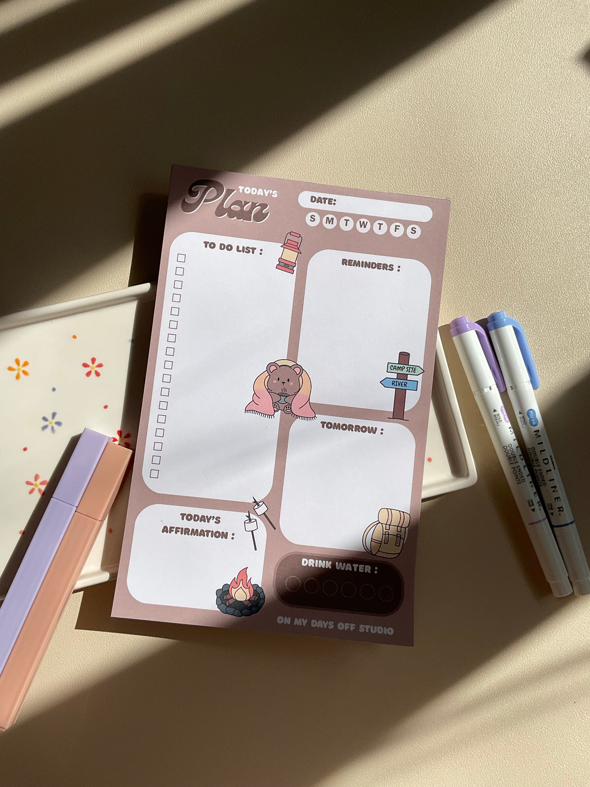 A daily planner notepad with a camp theme, featuring cute illustrations of a bear and camping related things. It has sections for a t do list, reminders, tomorrow list, today's affirmation and water in take tracker.