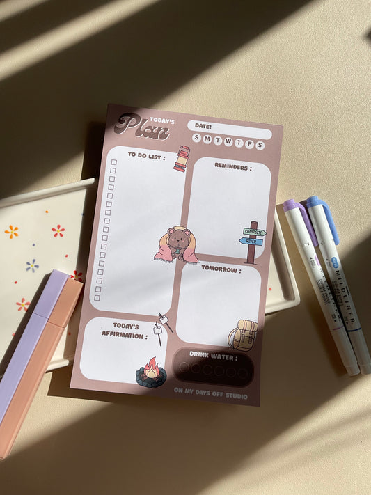 A daily planner notepad with a camp theme, featuring cute illustrations of a bear and camping related things. It has sections for a t do list, reminders, tomorrow list, today's affirmation and water in take tracker.
