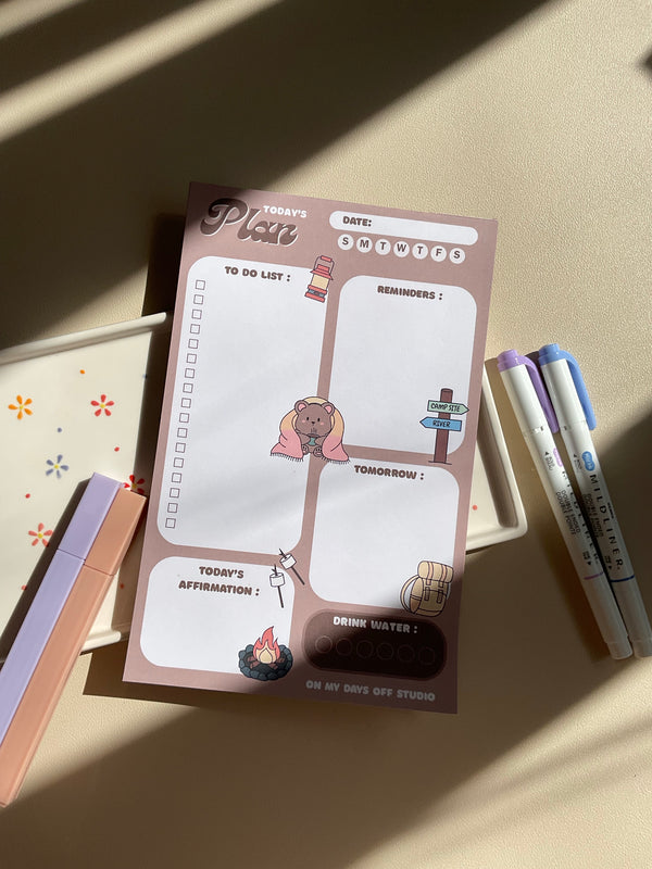 A daily planner notepad with a camp theme, featuring cute illustrations of a bear and camping related things. It has sections for a t do list, reminders, tomorrow list, today's affirmation and water in take tracker.