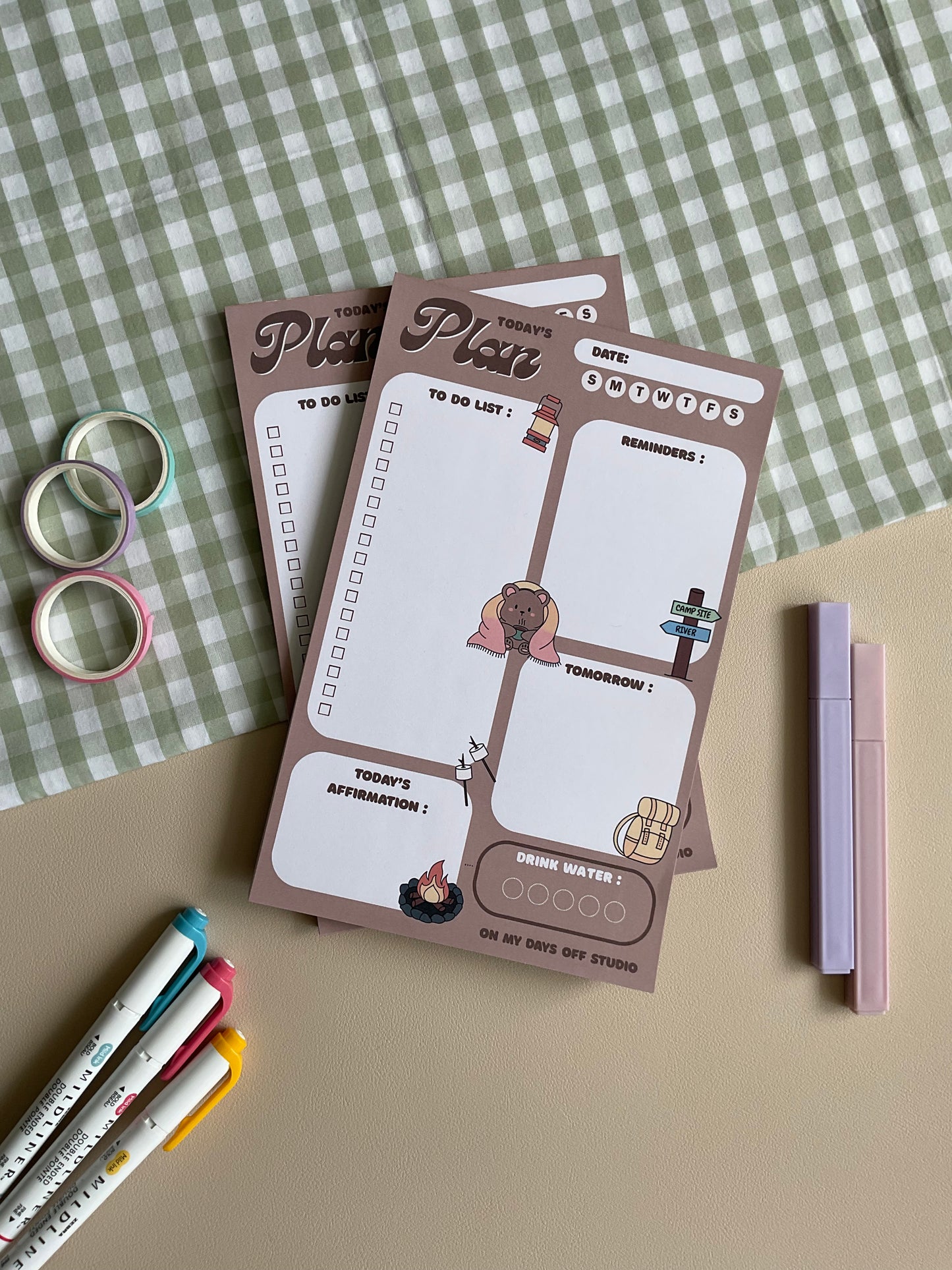 A daily planner notepad with a camp theme, featuring cute illustrations of a bear and camping related things. It has sections for a t do list, reminders, tomorrow list, today's affirmation and water in take tracker.