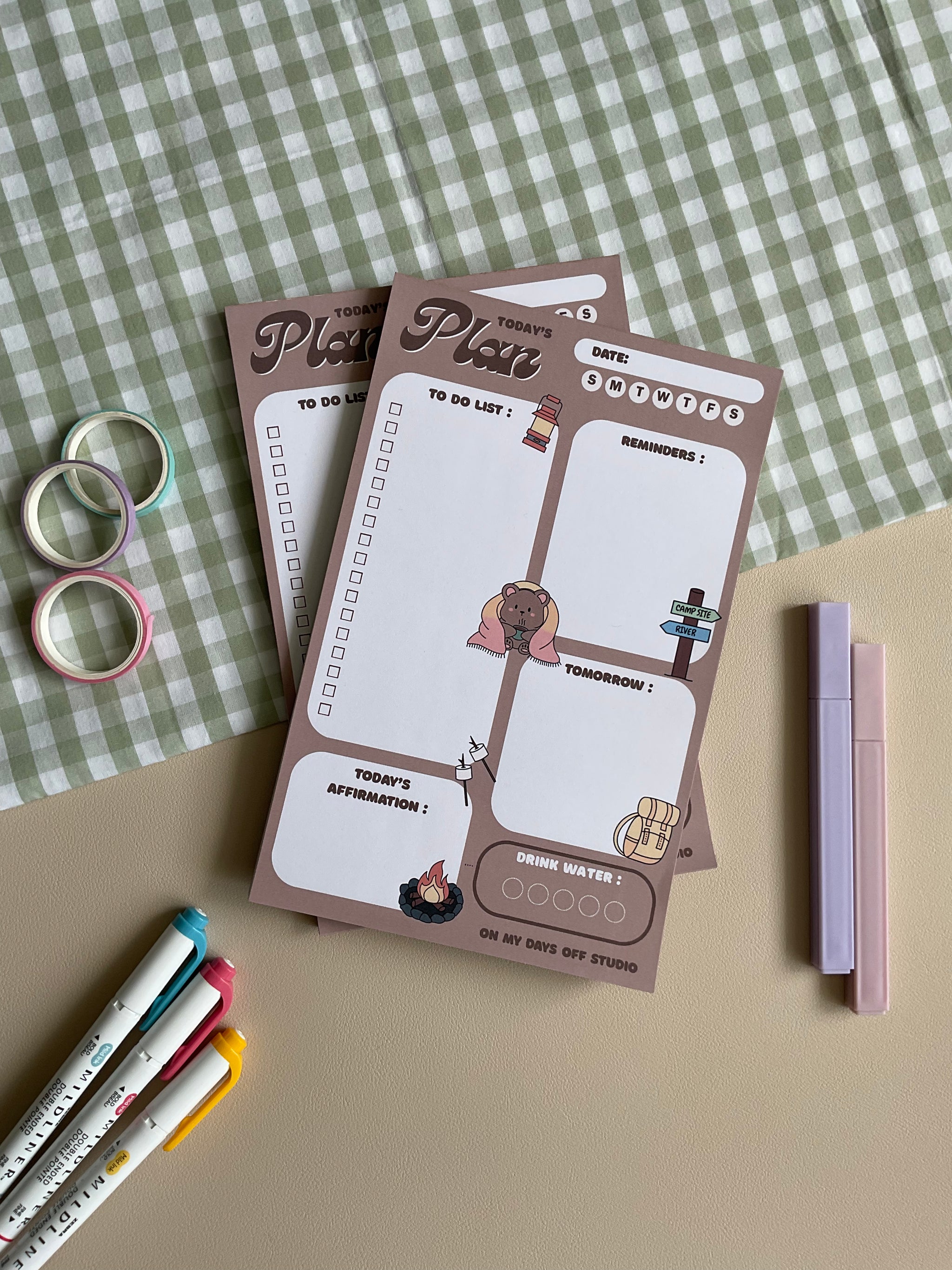 A daily planner notepad with a camp theme, featuring cute illustrations of a bear and camping related things. It has sections for a t do list, reminders, tomorrow list, today's affirmation and water in take tracker.