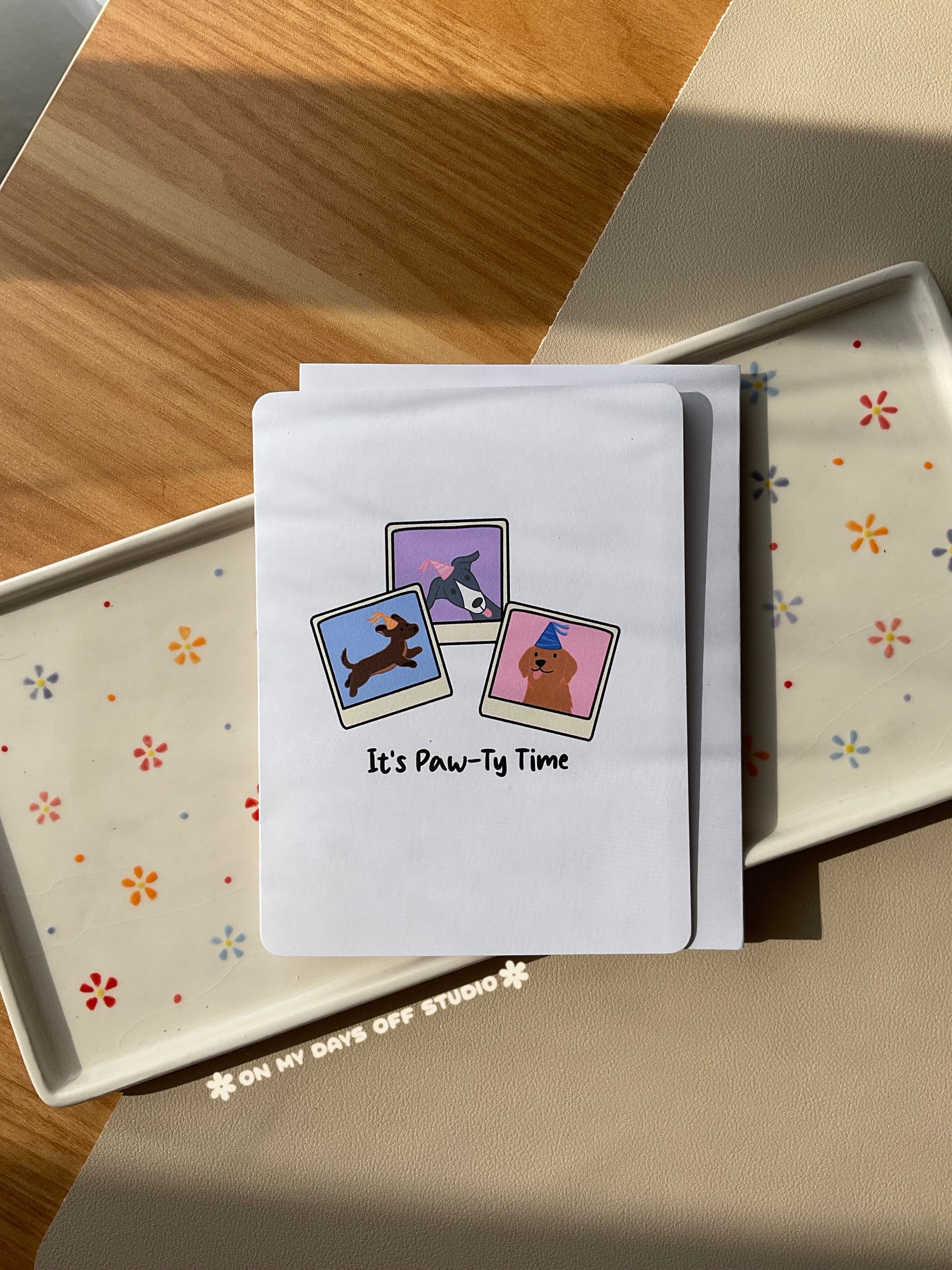 White greeting card with a cute illustration of 3 dogs in party hats & in a polaroids pictures. The card says "It's Paw-Ty Time". Blank interior for your personal handwritten message. 