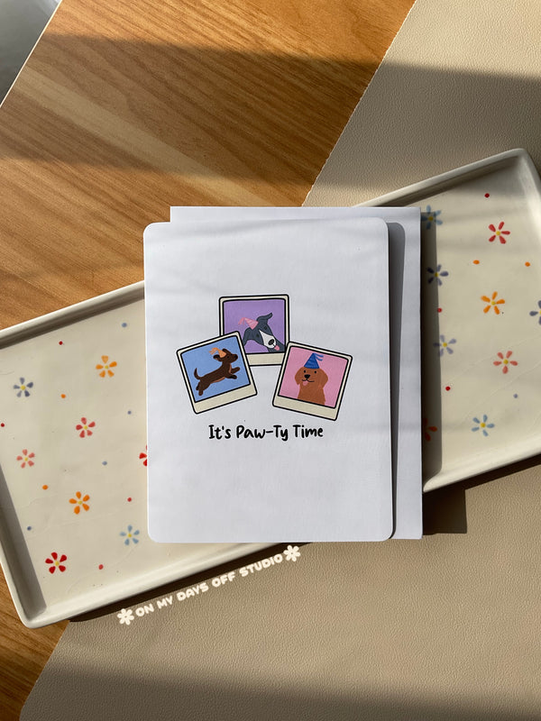 White greeting card with a cute illustration of 3 dogs in party hats & in a polaroids pictures. The card says "It's Paw-Ty Time". Blank interior for your personal handwritten message. 