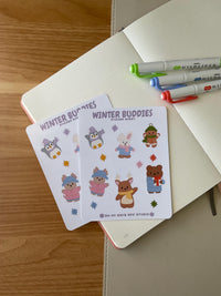 winter buddies sticker sheet front, featuring 6 illustrations of cute animals wearing winter clothes and accessories. This includes a penguin, bunny, gingerbread man, bear, deer and dog. Also, it includes 6 colorful stars. 